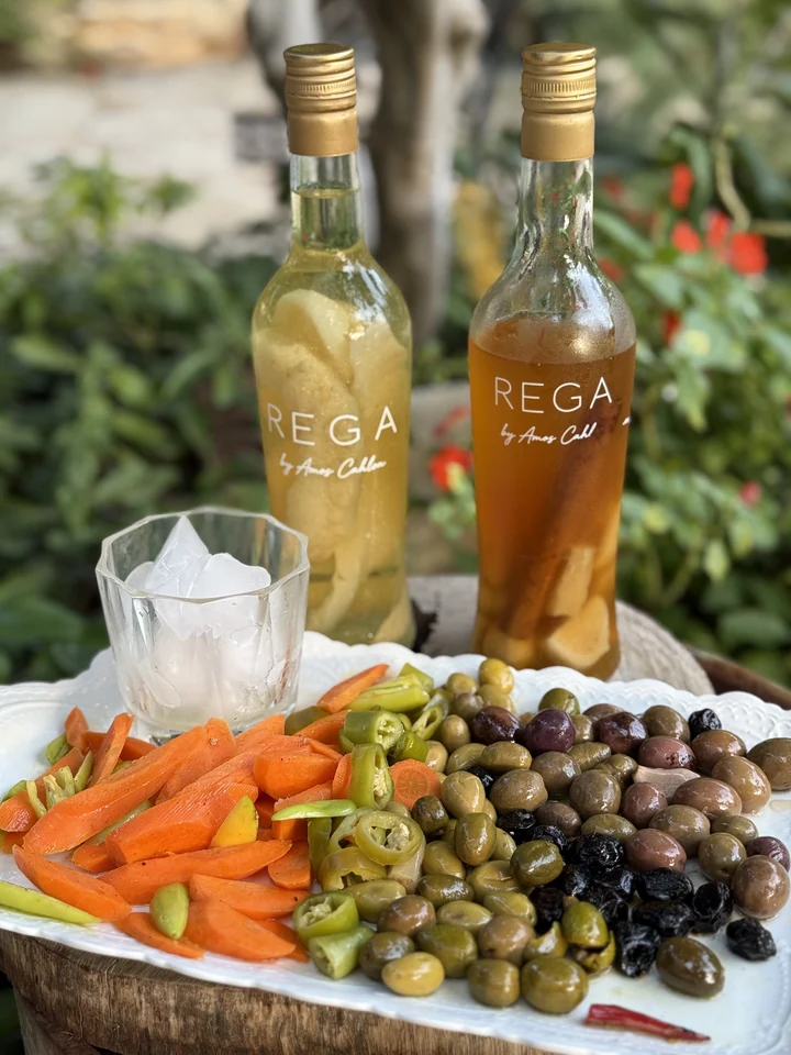 שני בקבוקי REGA צוננים בגווני צהוב וענבר, לצד כוס עם קרח, זיתים וירקות בגינה
