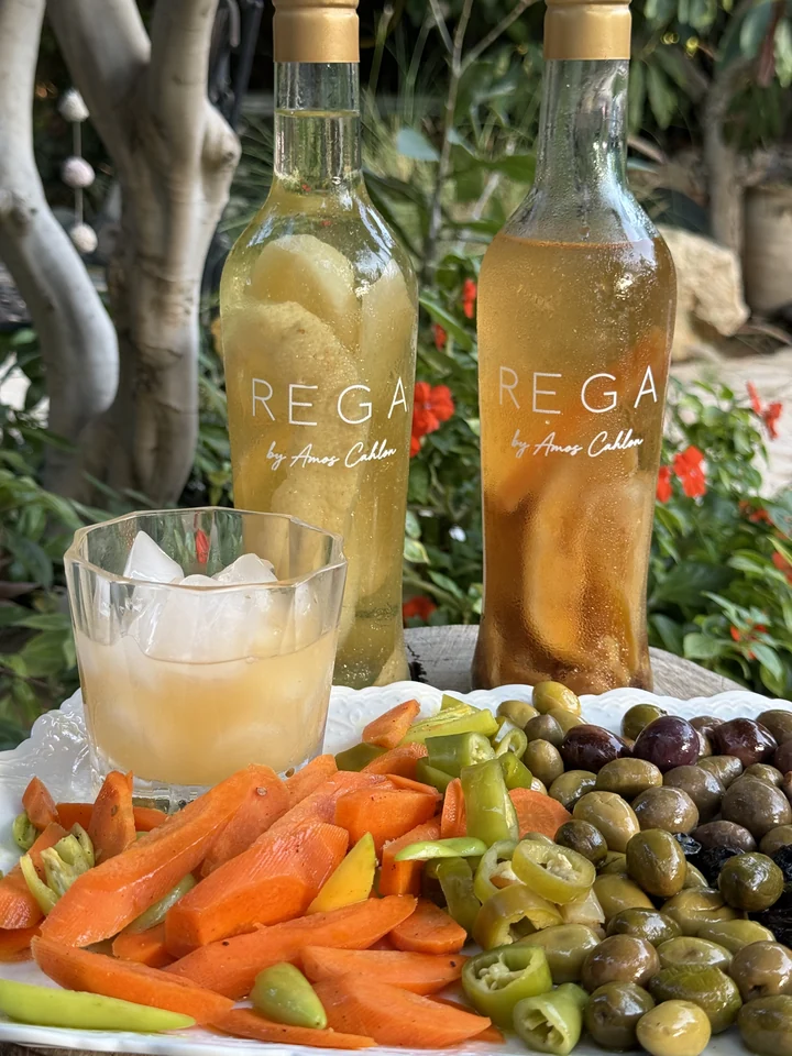 תקריב של שני בקבוקי REGA עם טיפות עיבוי וחתימת עמוס כחלון, לצד כוס, זיתים וירקות